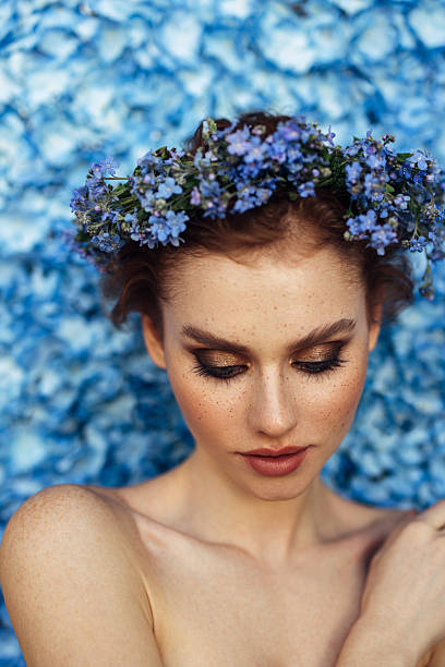 A striking woman with piercing blue eyes and delicate freckles poses gracefully against a sea of vivid blue flowers. A floral crown of the same hue sits atop her soft waves, accentuating her serene beauty. Her hand lightly touches her chin, adorned with a matching floral ring, adding to the harmony of the scene. The image radiates elegance, blending natural beauty with a touch of romance and artistry.