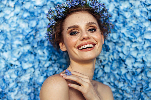 A striking woman with piercing blue eyes and delicate freckles poses gracefully against a sea of vivid blue flowers. A floral crown of the same hue sits atop her soft waves, accentuating her serene beauty. Her hand lightly touches her chin, adorned with a matching floral ring, adding to the harmony of the scene. The image radiates elegance, blending natural beauty with a touch of romance and artistry.