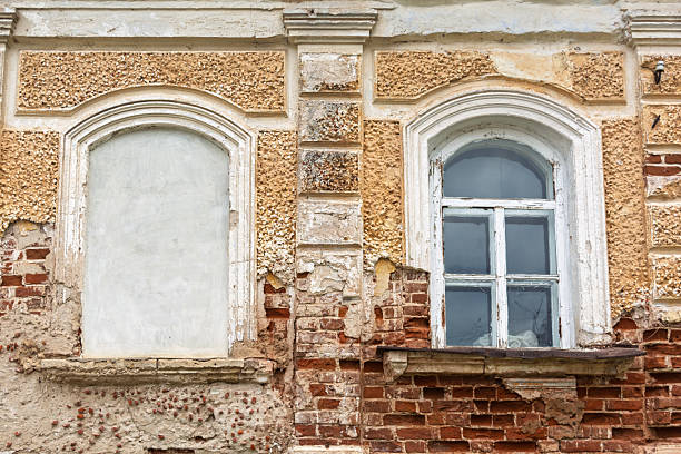Old brick wall with cracked plaster, one arched window bricked up, another with peeling paint, traces of time and neglect.