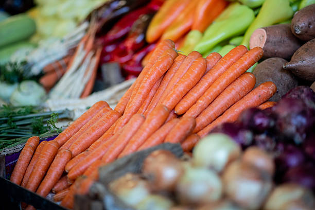 Fresh carrots take center stage in a vibrant market stall, surrounded by a colorful variety of other vegetables like peppers, greens, and root produce.