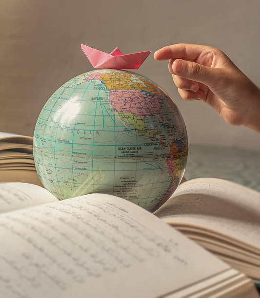 istock Global Journey: A hand places a pink paper boat on a globe model above open books 2226705582