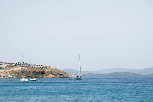 Two sailboats glide gently across calm sea waters, with a picturesque coastal hillside and distant mountains visible under clear skies. The scene captures a tranquil day at sea, ideal for sailing.