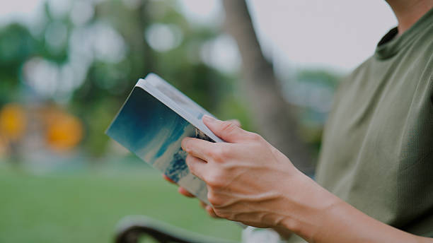 A close-up view of a young  man spending his morning to read a book while relaxing in a public park.