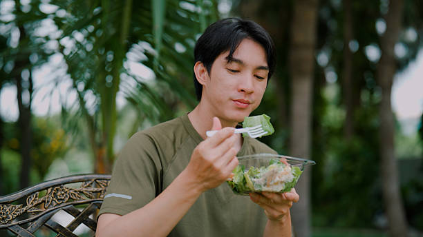 istock Young Asian man eating salad while sitting on a bench in a public park. 2226726398