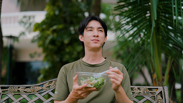 Young Asian man taking a moment to relax and enjoying green salad on a park bench.