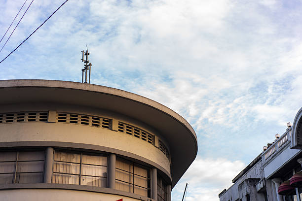 A partial view of a curved Art Deco-style building in Bandung with horizontal glass panel windows and minimalist architectural lines. The modernist roofline contrasts with the scattered clouds in the blue sky above, evoking a timeless urban atmosphere.