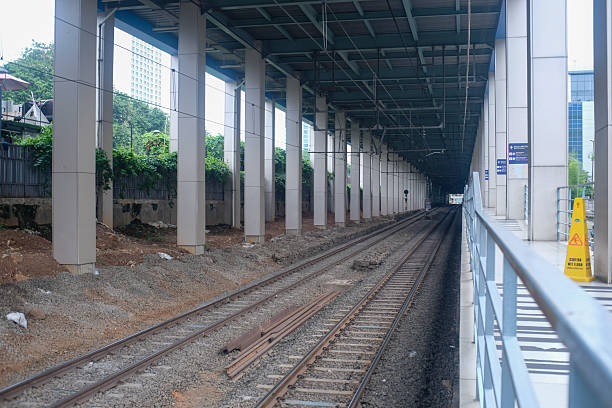 A railway track beneath an elevated platform in Jakarta, Indonesia, showcases modern urban infrastructure. The sheltered station features large support columns and a view of the long train tracks extending into the distance. The scene is calm, with construction materials visible alongside the tracks.