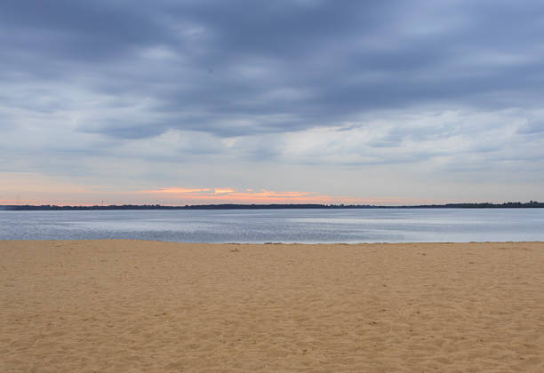 Sandy beach with a calm lake under moody clouds at sunset.