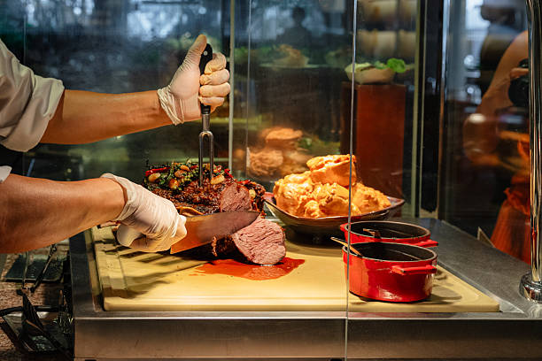 Chef slicing roast beef at a buffet carving station. The cooked meat is garnished with herbs and garlic, placed next to Yorkshire pudding and sauce pots. A professional food preparation scene suitable for restaurant, catering, or hospitality use.