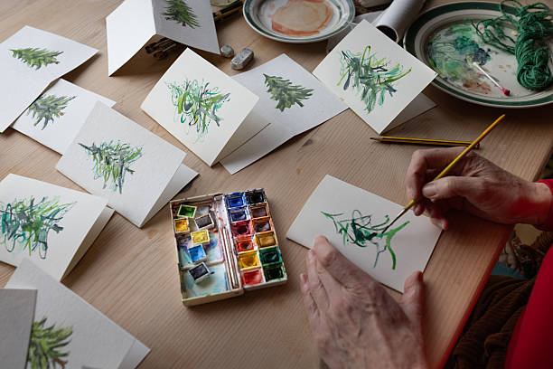 A medium over-the-shoulder shot of a mature woman sitting down at a wooden table in her family home. She is painting trees on homemade Christmas cards, using a selection of paintbrushes and watercolours.Videos are available similar to this scenario.