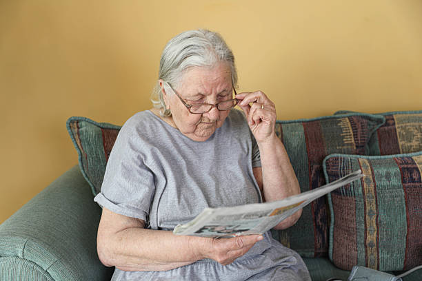Elderly Woman with Glasses Sitting on a Sofa, Concentrated While Reading a Newspaper. Indoor Scene Depicting Daily Routine, Information Access, and Senior Lifestyle at Home.
