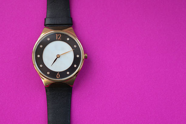 An elegant round wristwatch with a black and white dial and a gold-colored case, fastened with a black leather strap, is placed on a vibrant purple background.