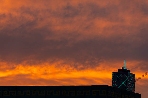 A vibrant orange sunset silhouettes a modern building with a pointed spire against dramatic clouds, with a textured foreground wall adding depth to this urban evening scene