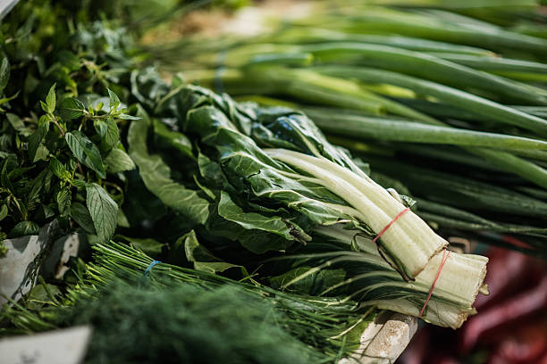 Neatly stacked bunches of chard, fresh herbs, spinach, and scallions fill the frame in a colorful close-up at a farmers market stall.