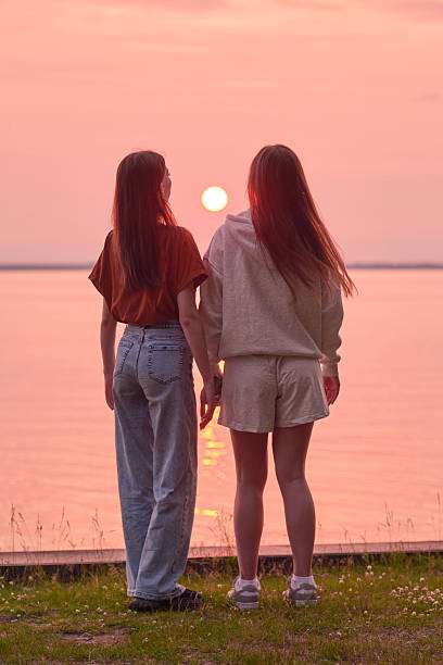 Young women, long brown hair, gaze at tranquil lake during sunset. Warm pink and orange hues cast over water and sky. Peaceful moment with natures beauty, grass foreground