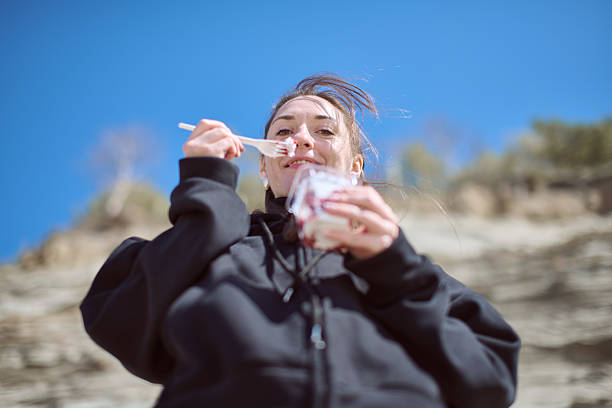 Young woman with brown hair relishes dessert outdoors. Dressed in black hoodie, she stands against cliff, serene and smiling. Crisp sunlight emphasizes joy and simplicity