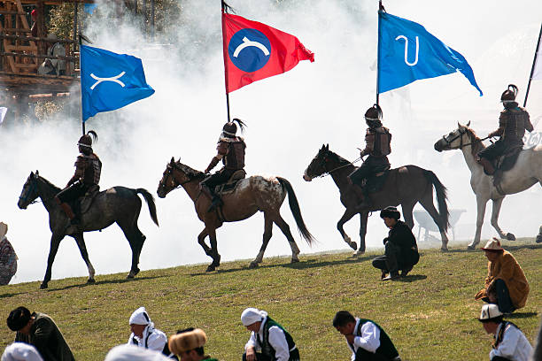 Three horsemen dressed in traditional warrior armor ride across a grassy field, each carrying a large flag—two red and one blue—during a performance at the World Nomad Games in Kyrgyzstan, 2016. White smoke drifts across the scene, partially obscuring the yurts and wooden structures in the background, creating a dramatic atmosphere. Seated and kneeling spectators in colorful traditional clothing watch from the foreground, highlighting the blend of cultural heritage and theatrical reenactment.