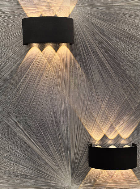 Two stylish wall-mounted lights illuminate a textured wallpaper surface, creating intricate patterns of light and shadow. The design enhances the ambiance of a contemporary space. Space for copy.