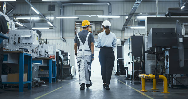 istock Diverse Female and Male Technicians Walking Through a Modern Factory, Discussing Work. Young Man Wearing a Yellow Hard Hat and Overalls, Manager Dressed in Business Attire. Footage From the Back 2226804305