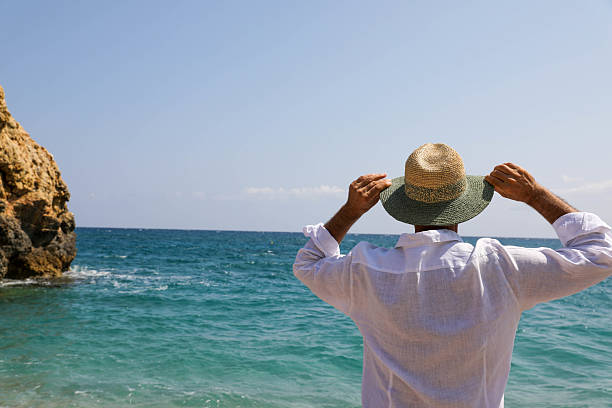 Mature Man in White Shirt and Straw Hat Gazing at the Ocean, Rear View Captured on a Sunny Summer Day by the Shore