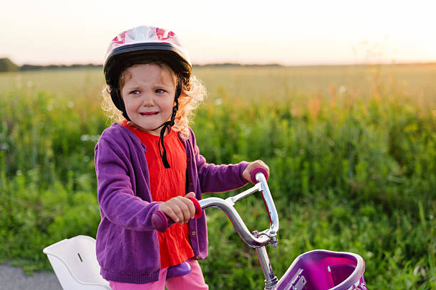 istock A small girl in a helmet looks distressed while holding her bicycle on a path, with a sunlit field in the background. 2226808487