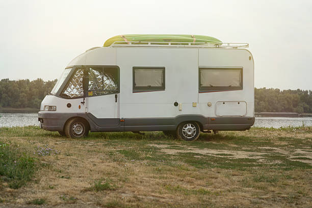 Side view of white camper van parked on grassy Danube riverbank, roof loaded with bright green kayak under warm evening light, symbolizing outdoor travel freedom and vanlife adventure.