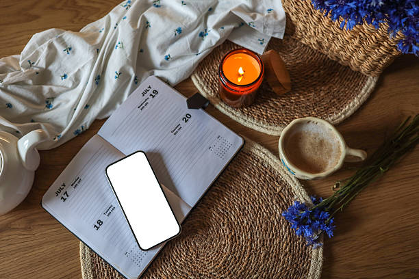 Flat lay with open planner, smartphone with blank screen, lit candle, coffee cup, and cornflowers on a wooden table. Cozy workspace with natural textures and warm light.