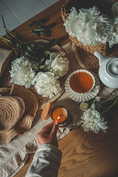 Hand lighting a candle in a cozy flat lay scene with tea, white peonies, straw hat, and an open book. Warm summer moment of relaxation and self-care in soft natural light.