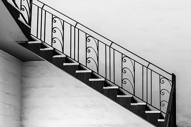 Black-and-white artistic photograph showcasing a decorative staircase with intricate wrought iron balustrade against plain walls, presenting a modern architectural design and a minimalist interior aesthetic.