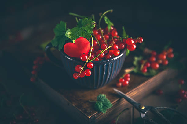 Freshly picked red currant berries and leaves are overflowing a green cup decorated with a red heart, sitting on a wooden box with gardening shears nearby