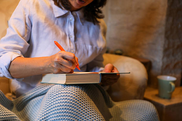 Senior woman sitting wrapped in a blanket, writing in a thick book or journal using an orange pen. Her hands are in the foreground, while the surrounding atmosphere radiates quiet home comfort. The photograph captures an intimate moment of focus, writing, or planning.