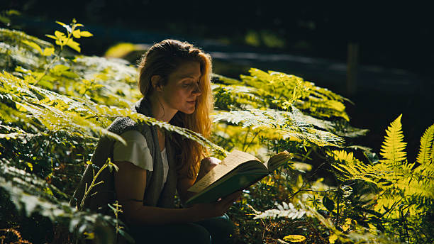 An american shot photo of a woman reads a book amidst lush ferns, basking in natural sunlight. The serene forest setting evokes a sense of tranquility and mindfulness, highlighting a harmonious connection with nature. Perfect for themes of relaxation and peaceful solitude.