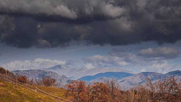 A dramatic scene showing dark storm clouds rolling over a vibrant autumn landscape, with rolling hills and forests in the foreground and snow-capped mountains in the distance. The image evokes a sense of awe and tranquility in the face of nature's power and beauty, making it perfect for themes of weather, travel, or natural beauty.