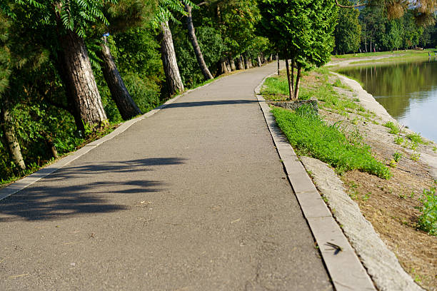 A well-maintained pathway runs alongside a calm river, bordered by greenery and tall trees. Sunlight filters through the leaves, creating a serene atmosphere.