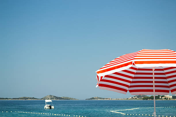 A vibrant red and white striped beach umbrella stands in the foreground against a backdrop of the calm blue sea and a clear, cloudless sky. In the distance, a small boat is visible on the water near some islands on the horizon. The composition has a minimalist feel with ample copy space, making it ideal for travel and vacation-themed designs. The image evokes a sense of peace, relaxation, and the perfect summer getaway.
