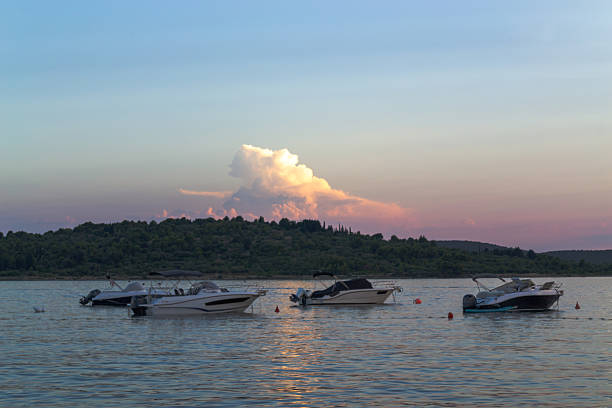 Several modern motorboats are anchored in a tranquil bay during a beautiful sunset. In the background, a lush green coastline sits under a colorful sky transitioning from blue to soft pink. A dramatic, illuminated cloud formation hovers above the hills, reflected in the calm water. The scene evokes a sense of peace, relaxation, and the perfect end to a summer day on the water.