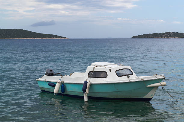 A small motorboat with a turquoise hull floats on the water near the shore of a historic coastal town, likely in Croatia. The background features traditional stone houses with red-tiled roofs situated on a peninsula, with green hills in the distance. The tranquil scene, set under a soft, cloudy sky, evokes the charm of a Mediterranean summer holiday and is perfect for travel-related content.