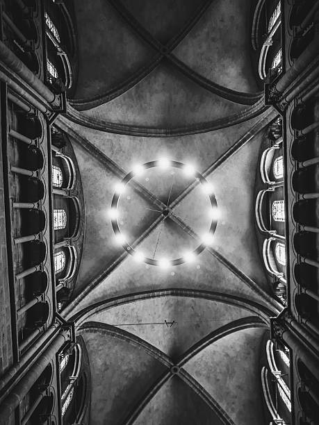 A black and white photograph looking up at a vaulted ceiling, likely in a Gothic or Romanesque cathedral or church. A circular light fixture with multiple bulbs is at the center, radiating light. Massive arches and ribs extend from the center, highlighting the grand and intricate architecture. Tall windows or niches are visible on the sides, emphasizing the structure's height. The interplay of light and shadow adds drama and texture.