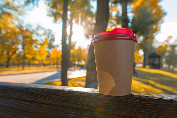 A brown paper coffee cup with a red lid rests on a wooden railing in a sun-drenched autumn park with golden trees in the background
