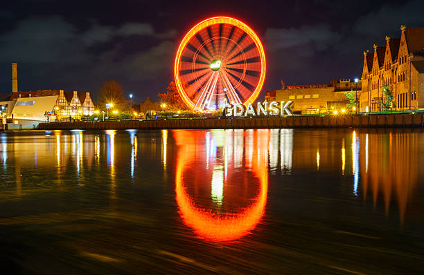 Illuminated ferris wheel and Gdask sign reflecting in Motawa River at night, with vibrant lights and historic buildings. Poland.