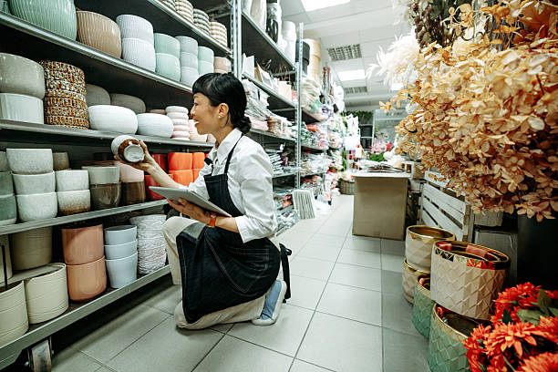 Woman in a plant store, focused on a digital tablet as she checks ceramic pots on display. She is reviewing inventory or completing a checklist for store organization or stock planning.