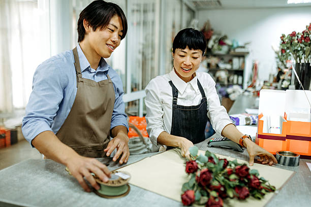 Two florist colleagues are working together in a flower shop, carefully arranging fresh red roses into a beautiful bouquet.