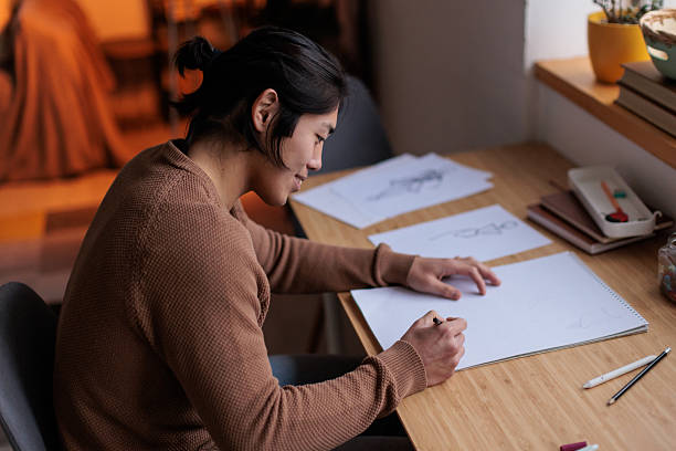 Young Japanese man expresses his creativity by drawing sketches on paper while sitting at his home desk.