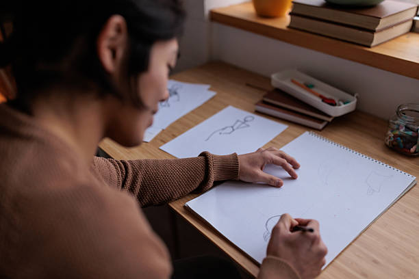 Young Japanese man expresses his creativity by drawing sketches on paper while sitting at his home desk.