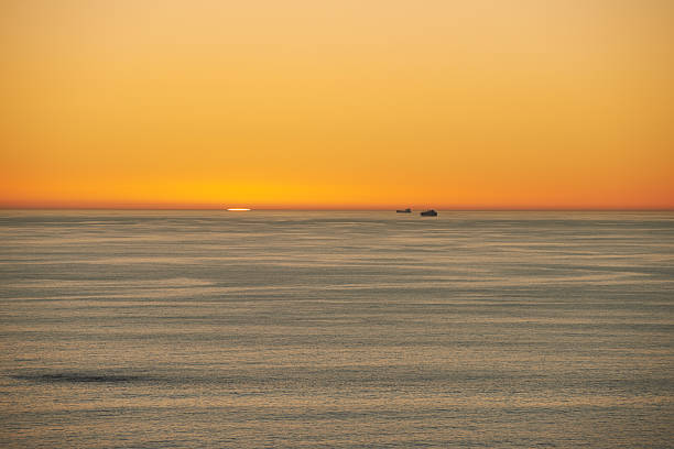 Golden hues adorn the calm evening sea as the sun sets, casting warm light across the waves. Far in the distance, two ships are silhouetted against the serene horizon.