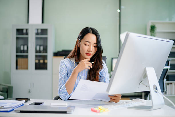 istock Asian businesswoman stands at her desk writing on a clipboard while analyzing data charts in a bright, modern office 2226893608