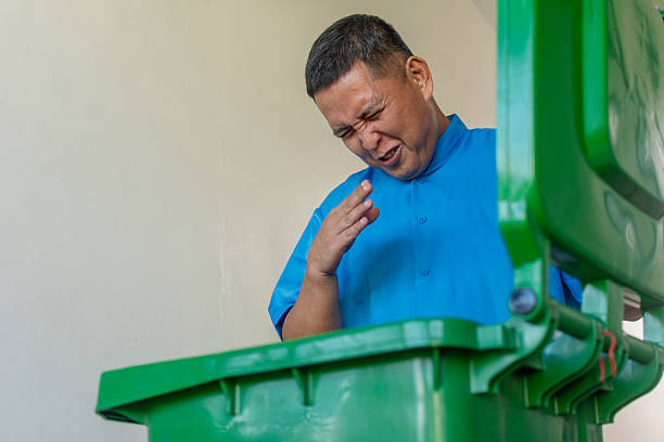 A middle aged asian janitor is disgusted from the bad smell emanating inside a large plastic mobile trash bin.