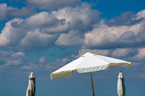 A single man on a sandy beach, wearing a protective face mask and sunglasses sits next to an  empty deck chair, near the parasol and rubber duck inflatable,