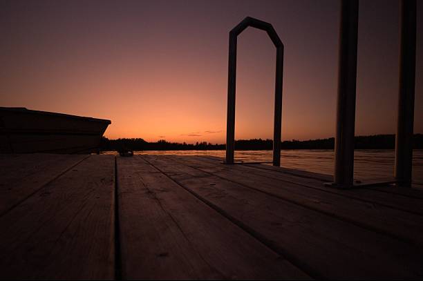 A serene sunset view from a wooden dock overlooking a calm lake with a boat and distant trees, creating a tranquil atmosphere.