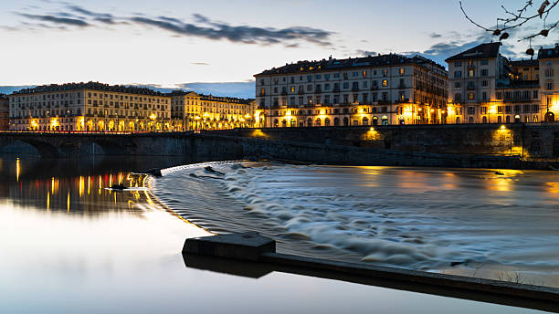 At the blue hour, darkness slowly descends on the streets of Turin, highlighting the light trails of cars driving along Piazza Vittorio Veneto and Vittorio Emanuele I Bridge. The sky is clearing from the clouds, reflected in the waters of Po river. Magic is in the air.
